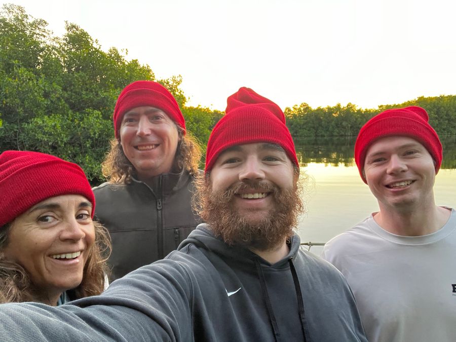 Four people in matching red beanies by a mangrove creek